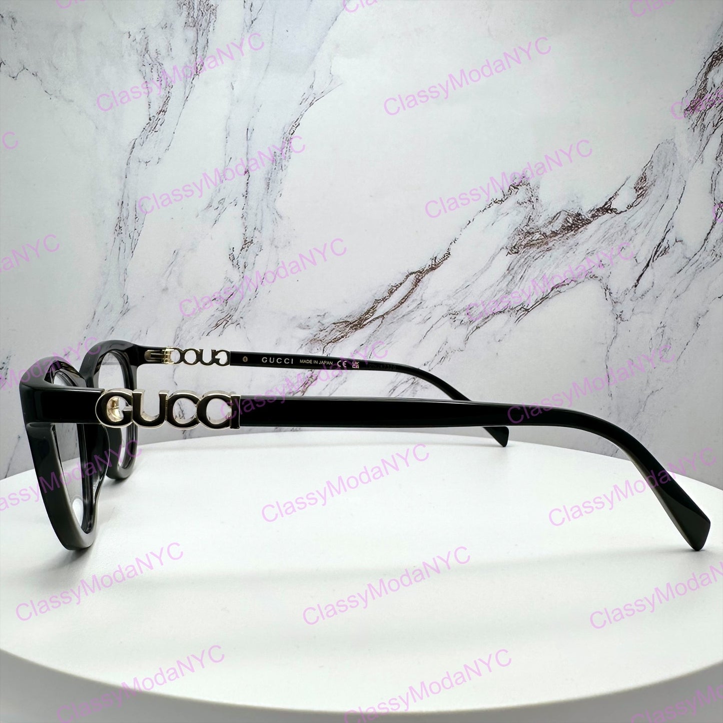 Gucci black acetate square eyeglasses with gold-tone metal frames and interlocking GG logo details on the temples, displayed on a white round pedestal against a white marble-patterned background.